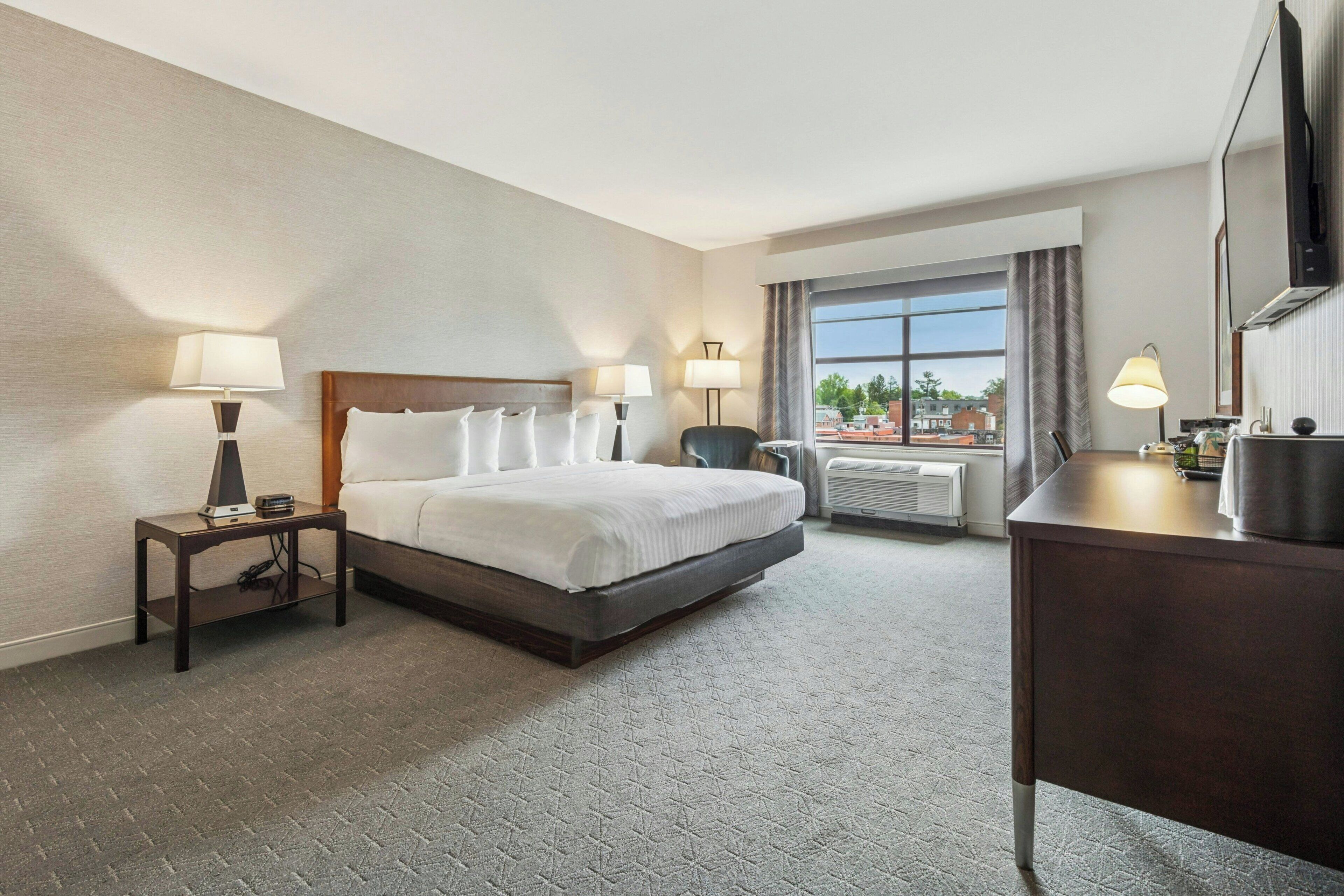 A hotel room with a bed, bedside lamps, a desk, a chair, and a window with a view.