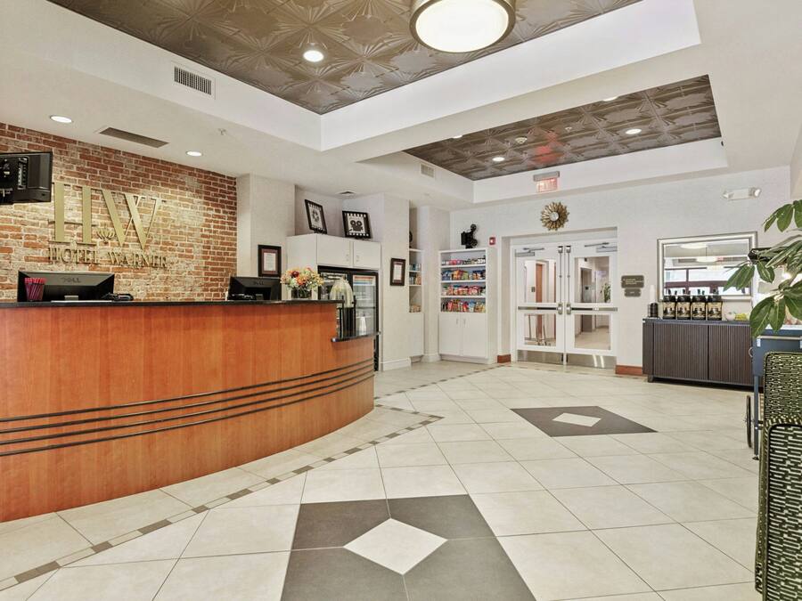 Zona de recepción de un hotel con pared de ladrillo, mostrador de recepción, televisor, cuadros enmarcados, máquina de café, planta y quiosco de autoservicio.