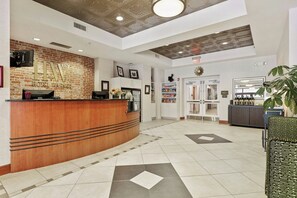 Zona de recepción de un hotel con pared de ladrillo, mostrador de recepción, televisor, cuadros enmarcados, máquina de café, planta y quiosco de autoservicio.