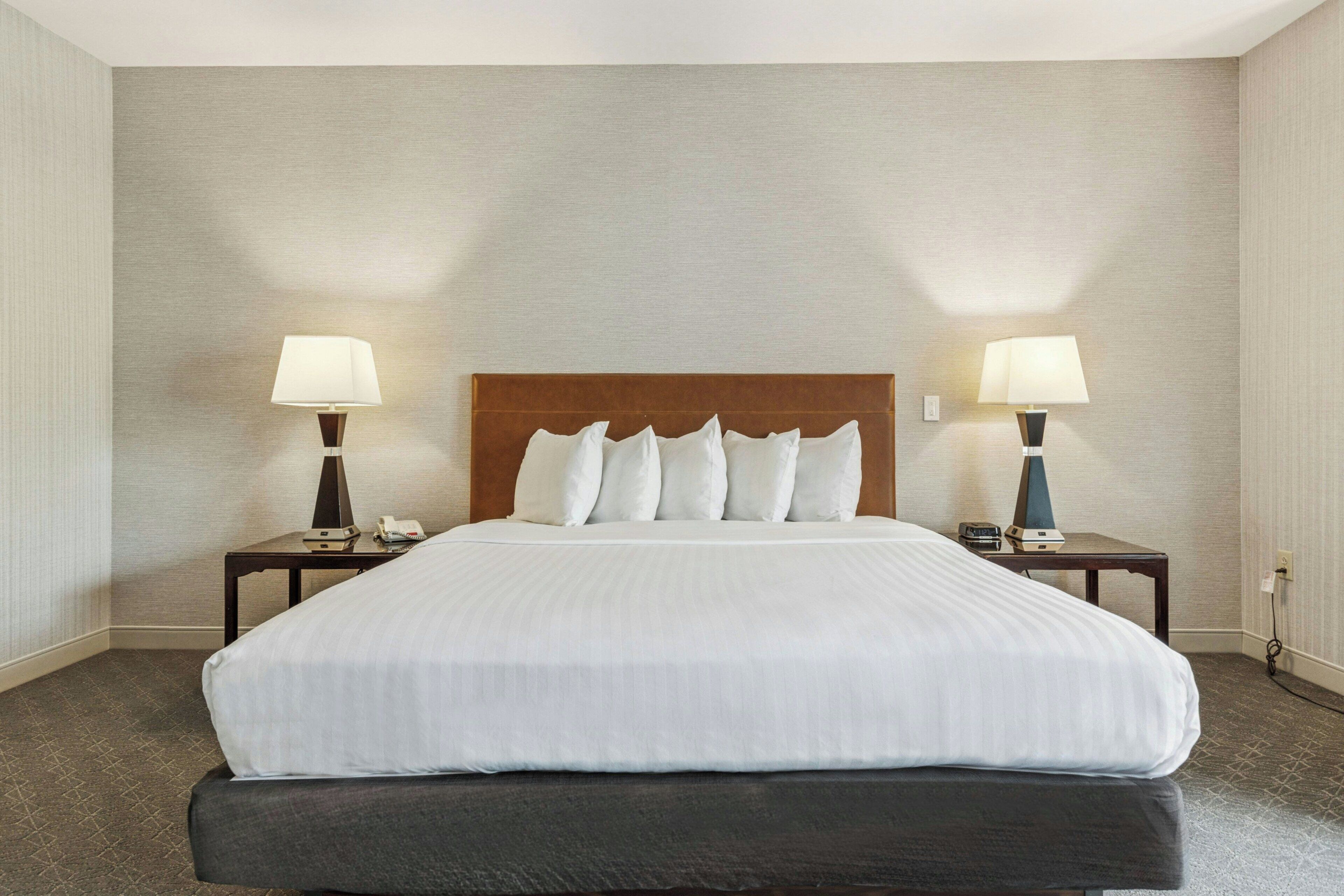Una habitación de hotel con una cama grande, dos mesitas de noche con lámparas y una pared lisa.