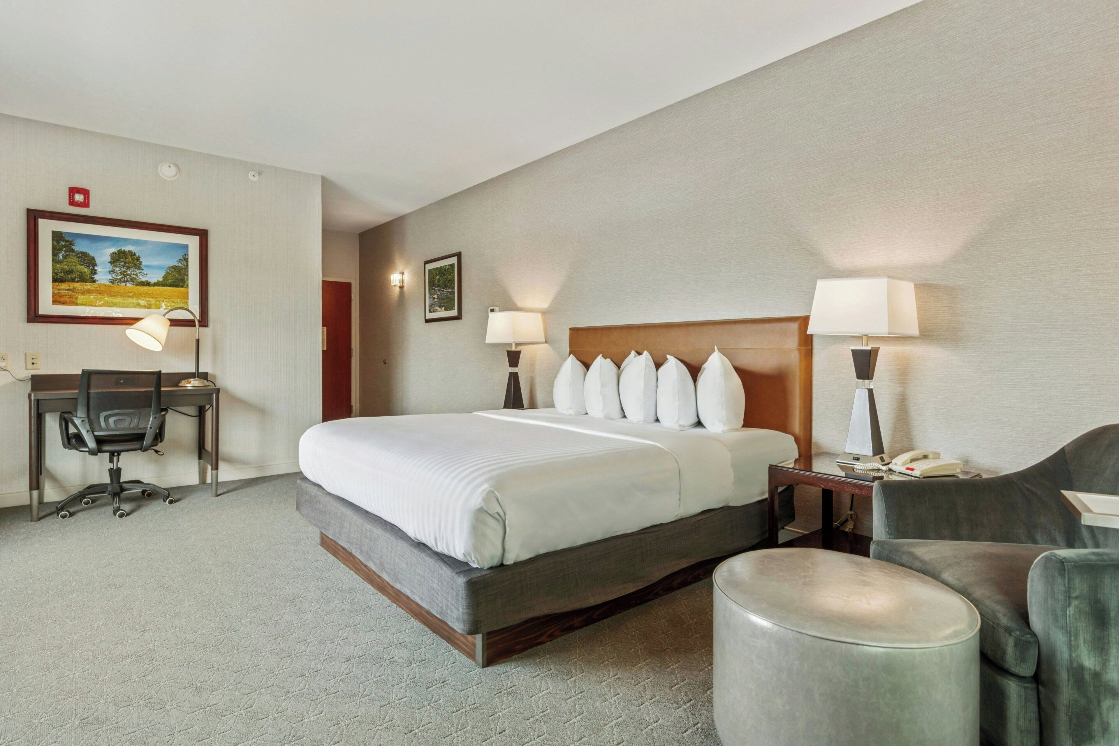 A hotel room with a bed, desk, chair, and ottoman.