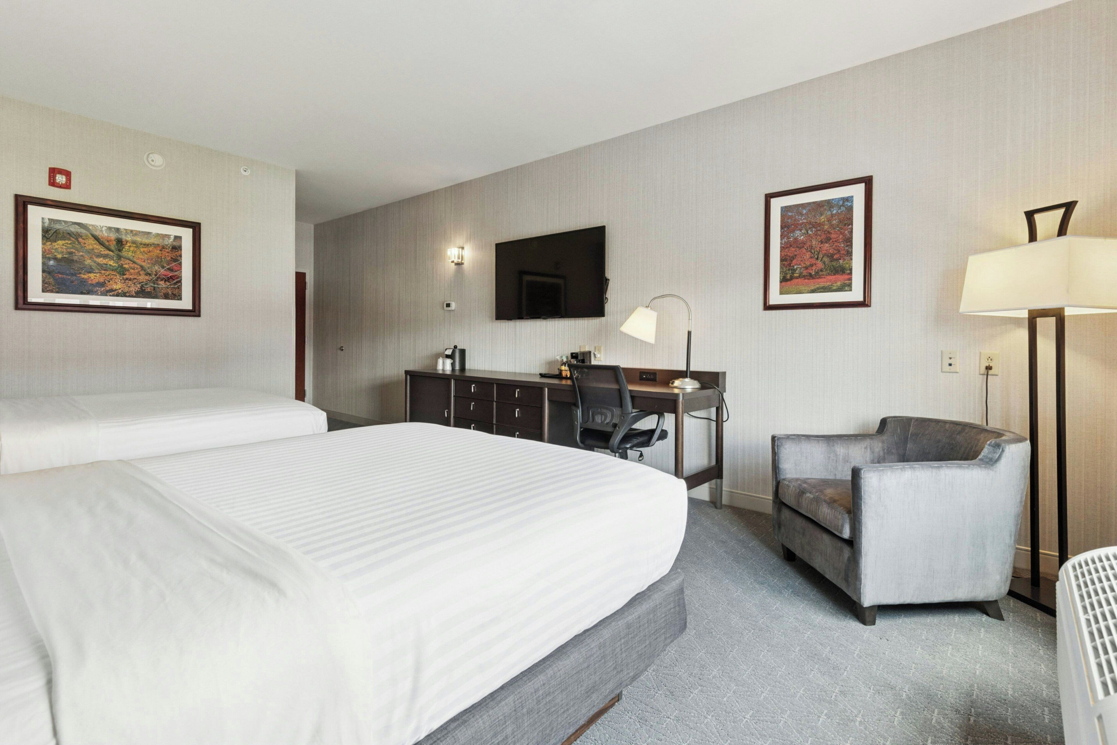 A hotel room with two beds, a desk with a chair, a TV, and a lamp.