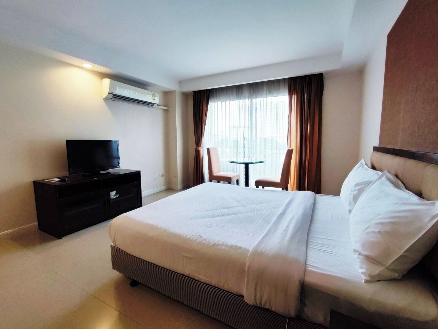 A hotel room with a large bed, a TV on a stand, a small table with two chairs, and a window with curtains.