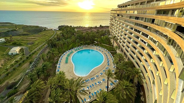 A hotel with a pool surrounded by palm trees and a view of the ocean at sunset. - Club Hotel Marazul (Adeje)