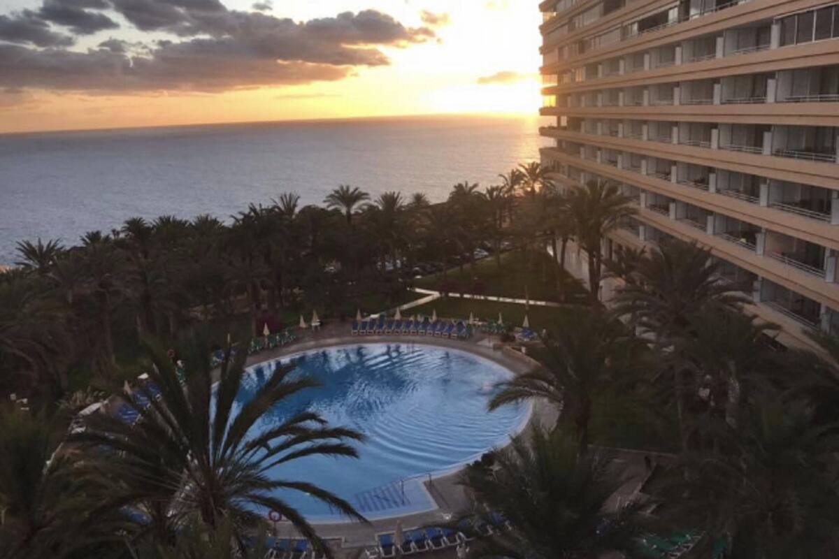 A resort pool with a view of the ocean at sunset, surrounded by palm trees and a multi-story building.