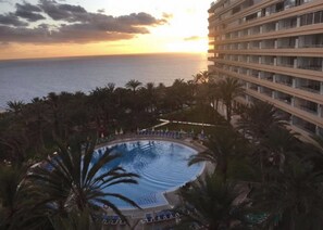 Zwembad van een resort met uitzicht op de oceaan bij zonsondergang, omringd door palmbomen en een meerlaags gebouw.