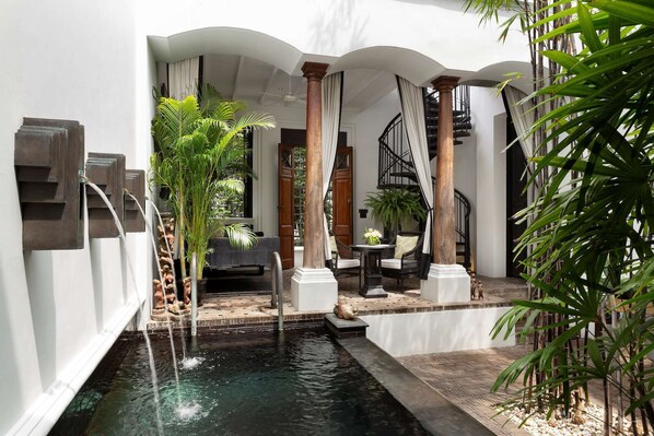 A serene indoor garden with a central water feature, stone steps, and a seating area with a table and chairs.