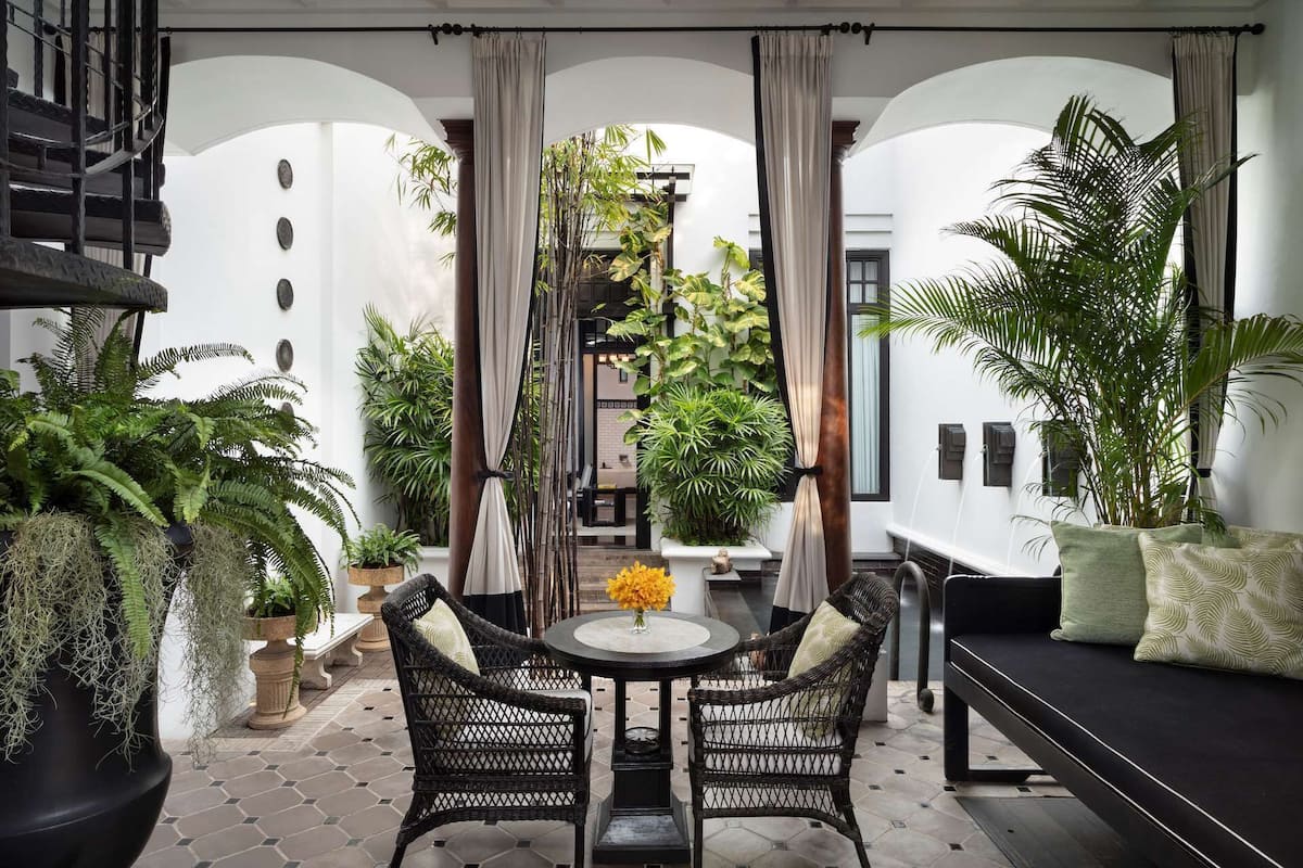 Un espacio exterior acogedor con sillas de mimbre y un sofá negro, rodeado de plantas verdes exuberantes y un suelo de baldosas.