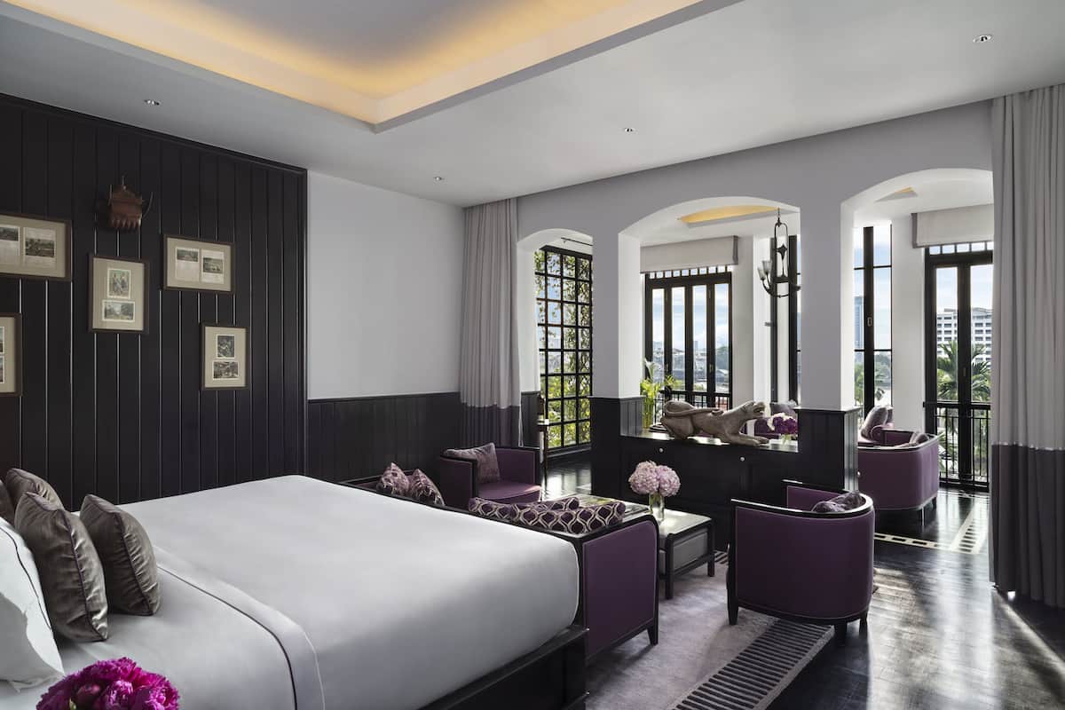 Una habitación de hotel moderna con una cama grande, un área de descanso con sillas moradas y vistas a la ciudad a través de amplios ventanales.