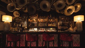 A bar with multiple hanging brass instruments, a well-stocked liquor shelf, and red upholstered bar stools.