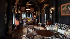 A cozy, dimly lit room with wooden walls and a high ceiling. There is a round table with chairs, a sofa with a zebra print cover, and various framed pictures and decorations on the walls.