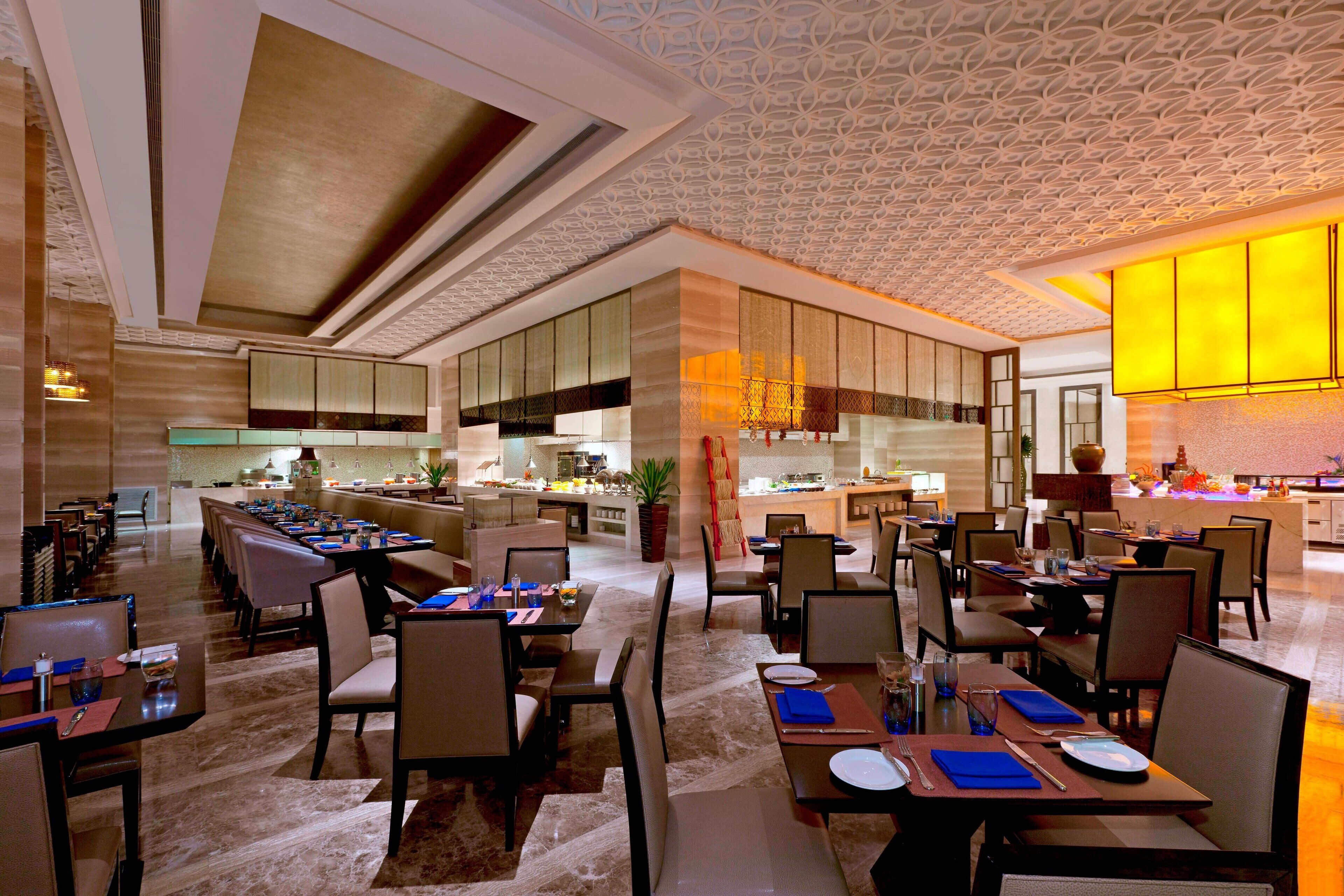 A spacious hotel dining area with multiple tables and chairs, a kitchen in the background, and a decorative ceiling.