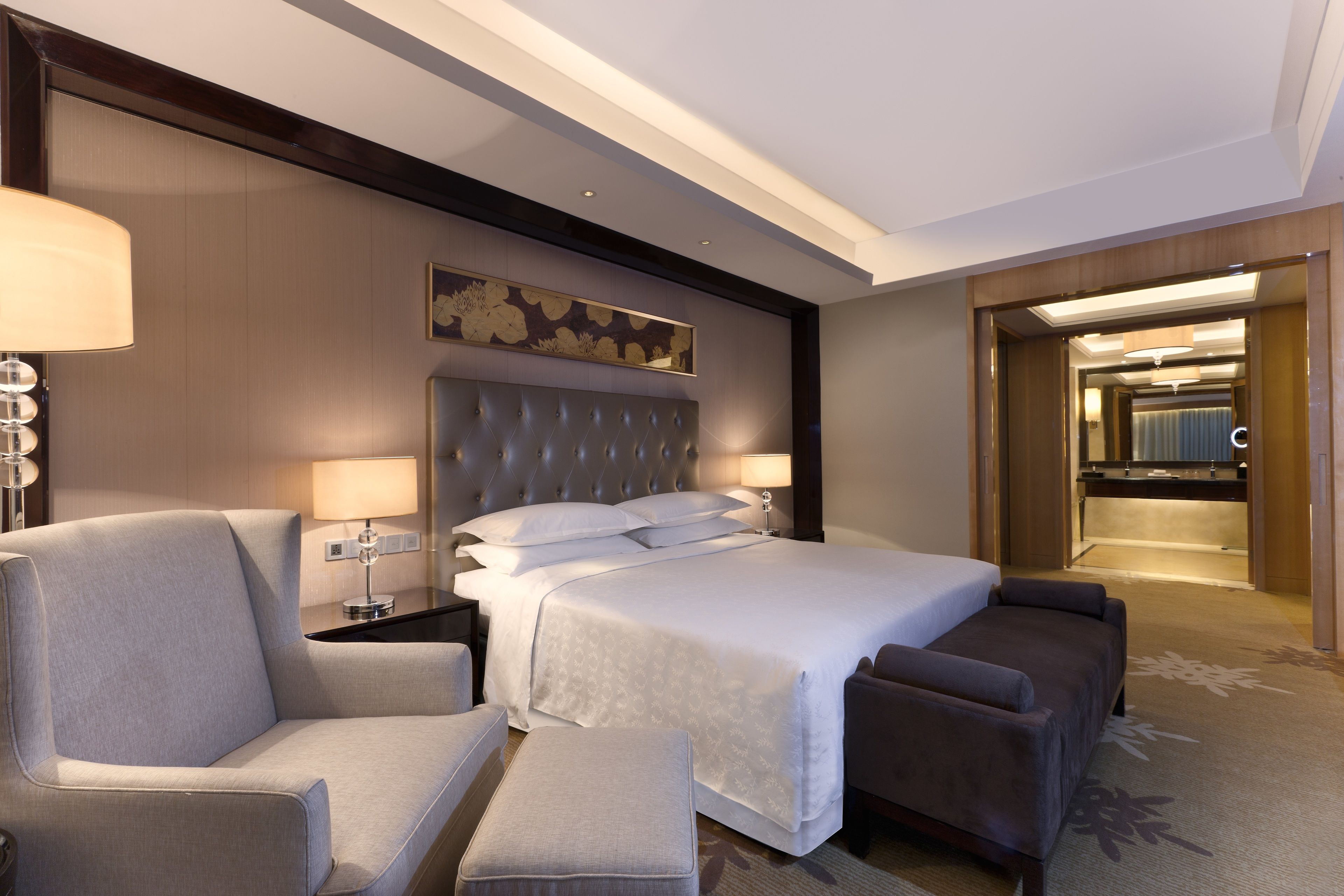 Une chambre d’hôtel moderne dotée d’un grand lit, d’un coin salon avec un canapé et un fauteuil, d’une table de chevet avec une lampe, et d’un couloir menant à la salle de bain.
