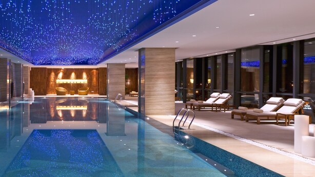 Een modern binnenzwembad met een spiegelend wateroppervlak, lounge stoelen en een plafond versierd met hangende lampen.