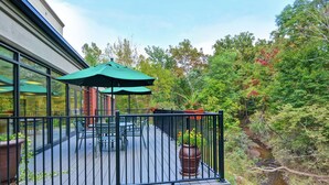 Une terrasse dotée de mobilier d’extérieur et de parasols, entourée d’une rambarde en métal et offrant une vue sur une zone boisée.