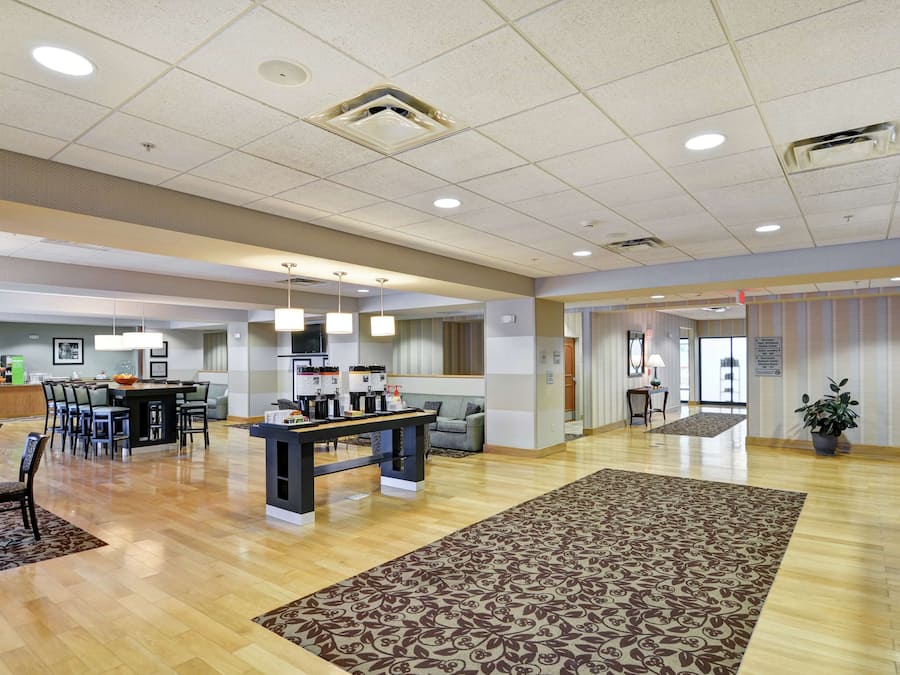 A spacious lobby with a wooden floor, a central coffee station, seating areas, and a patterned rug.