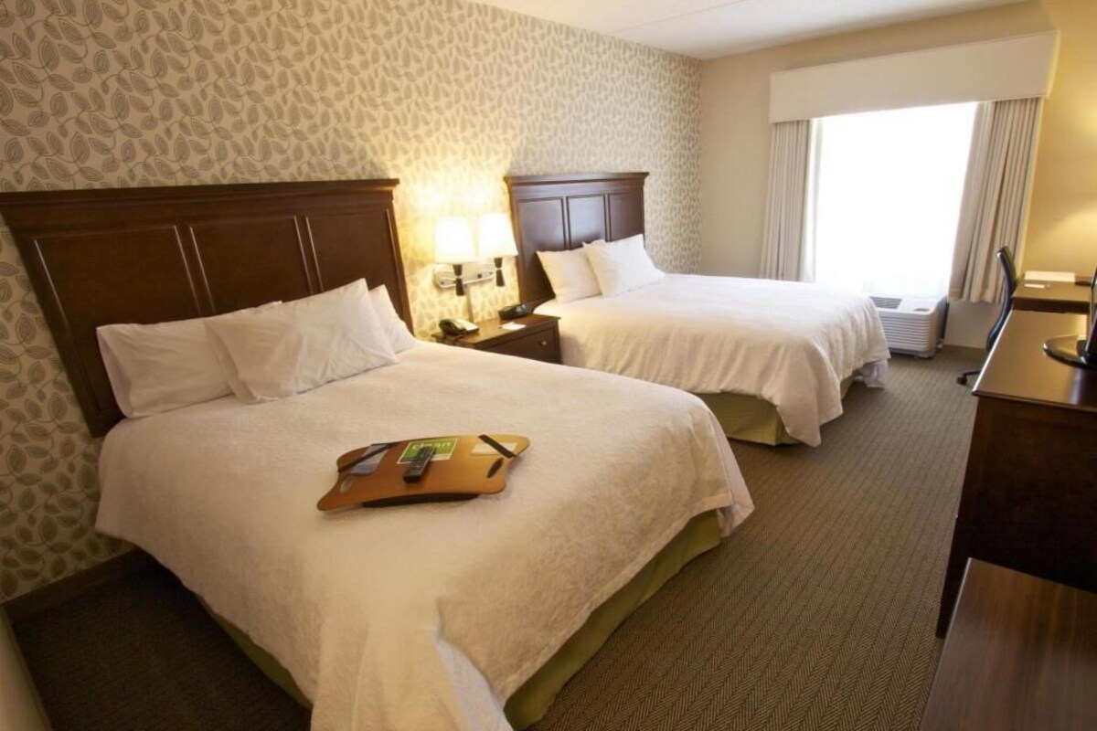 A hotel room with two beds, a desk, a chair, a nightstand, and a window with curtains.