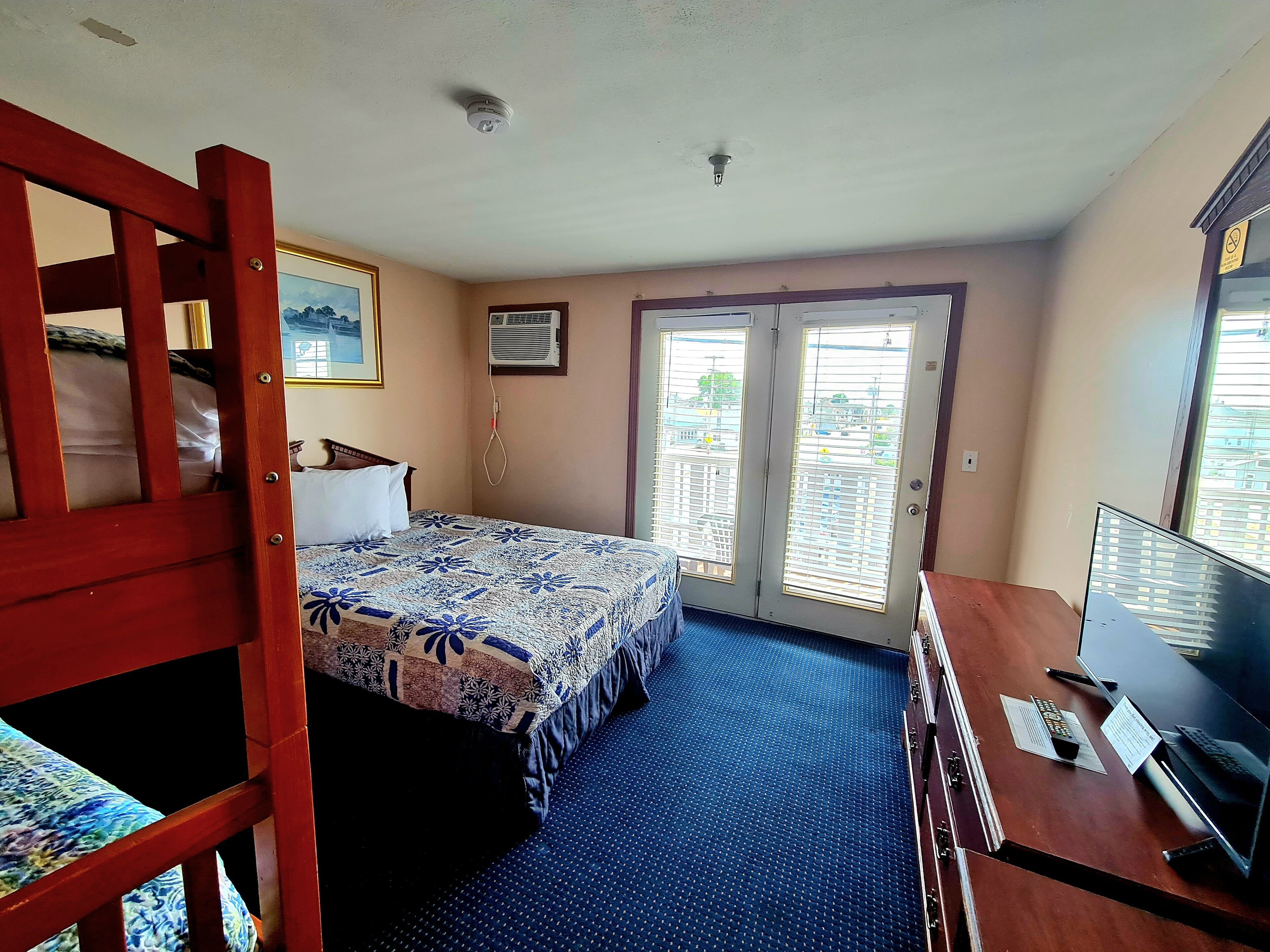 A room with a bed, a desk with a computer, a window with blinds, and a door.