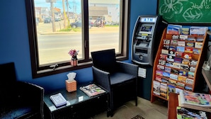A room with a window overlooking a street, a small table with a tissue box and magazines, a chair, and an ATM. - Regal Inn (Hampton)