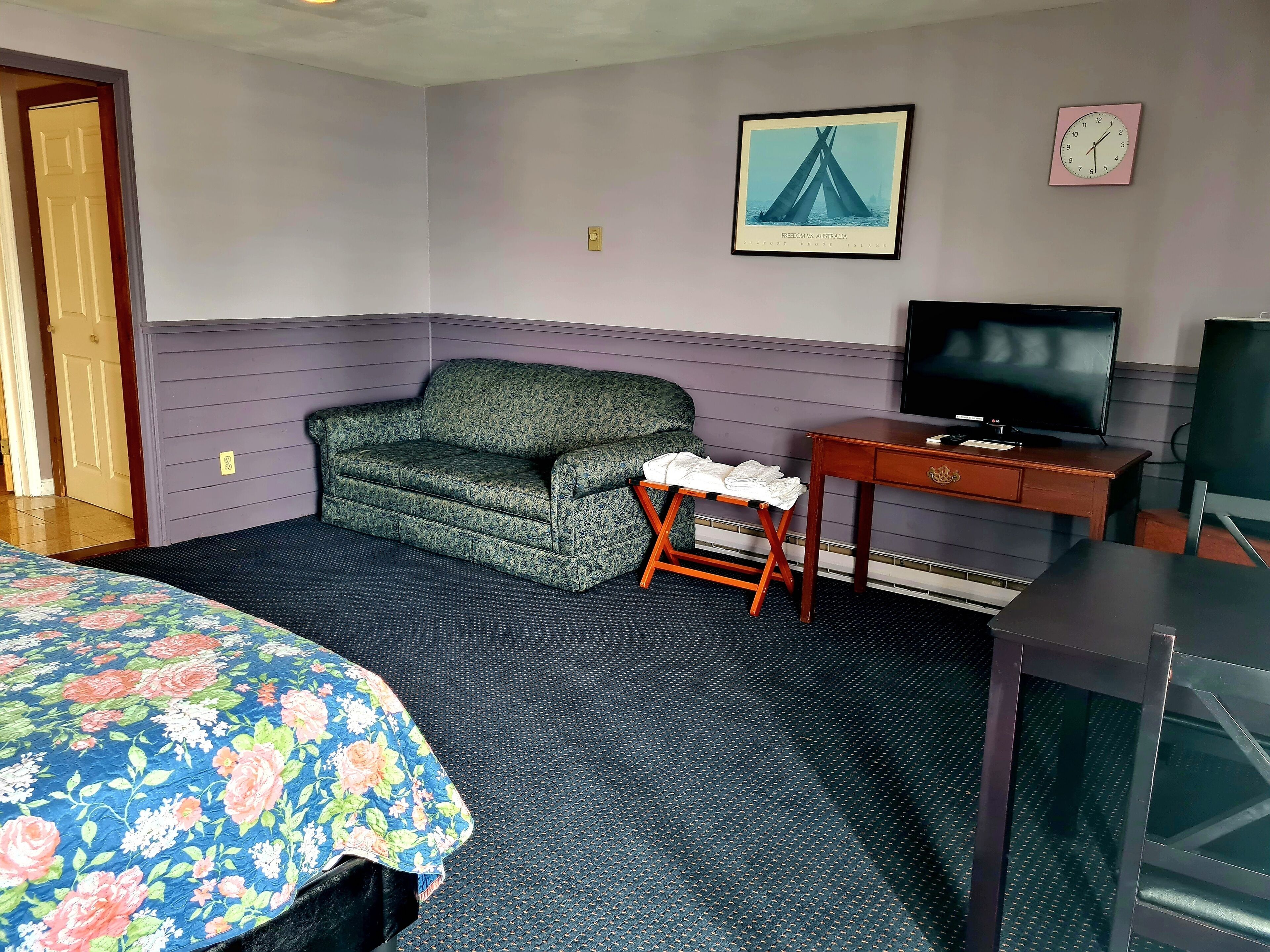 A room with a bed, a green patterned sofa, a small wooden table with a TV, and a framed picture on the wall.