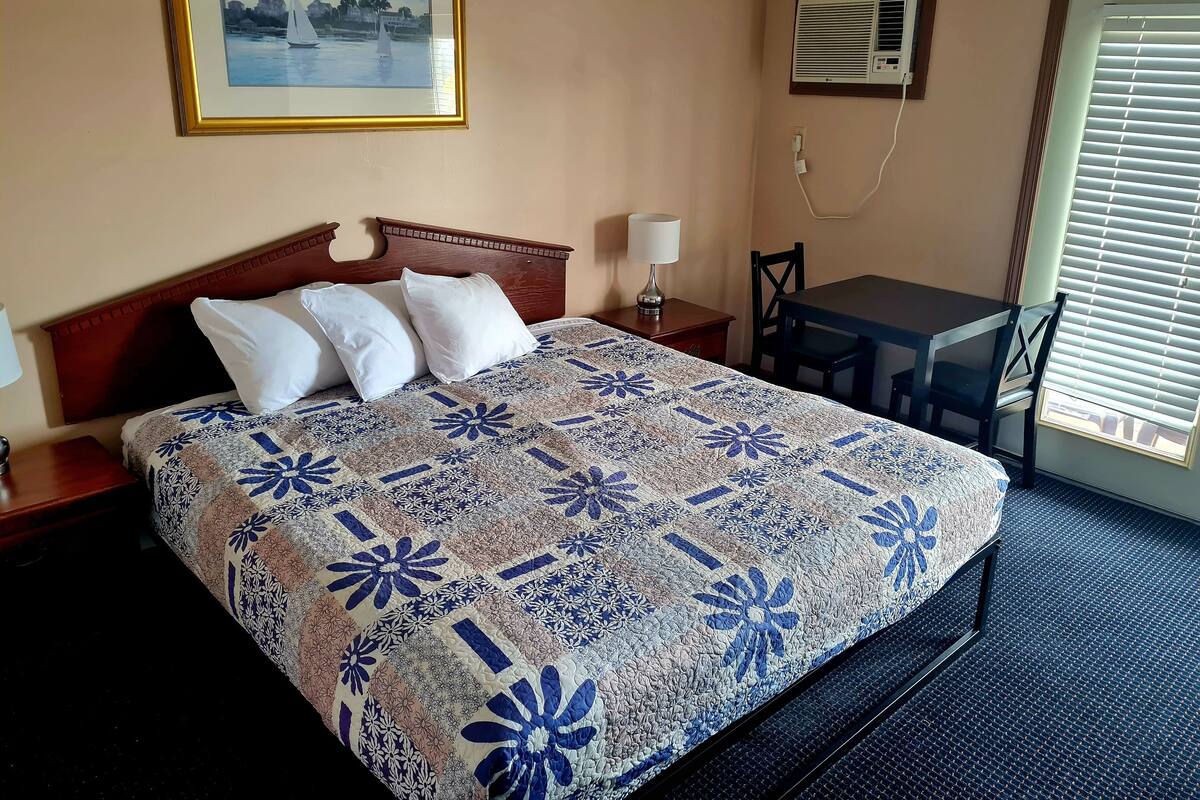 A hotel room with a bed, a desk, and a chair. There is a painting on the wall and an air conditioning unit.