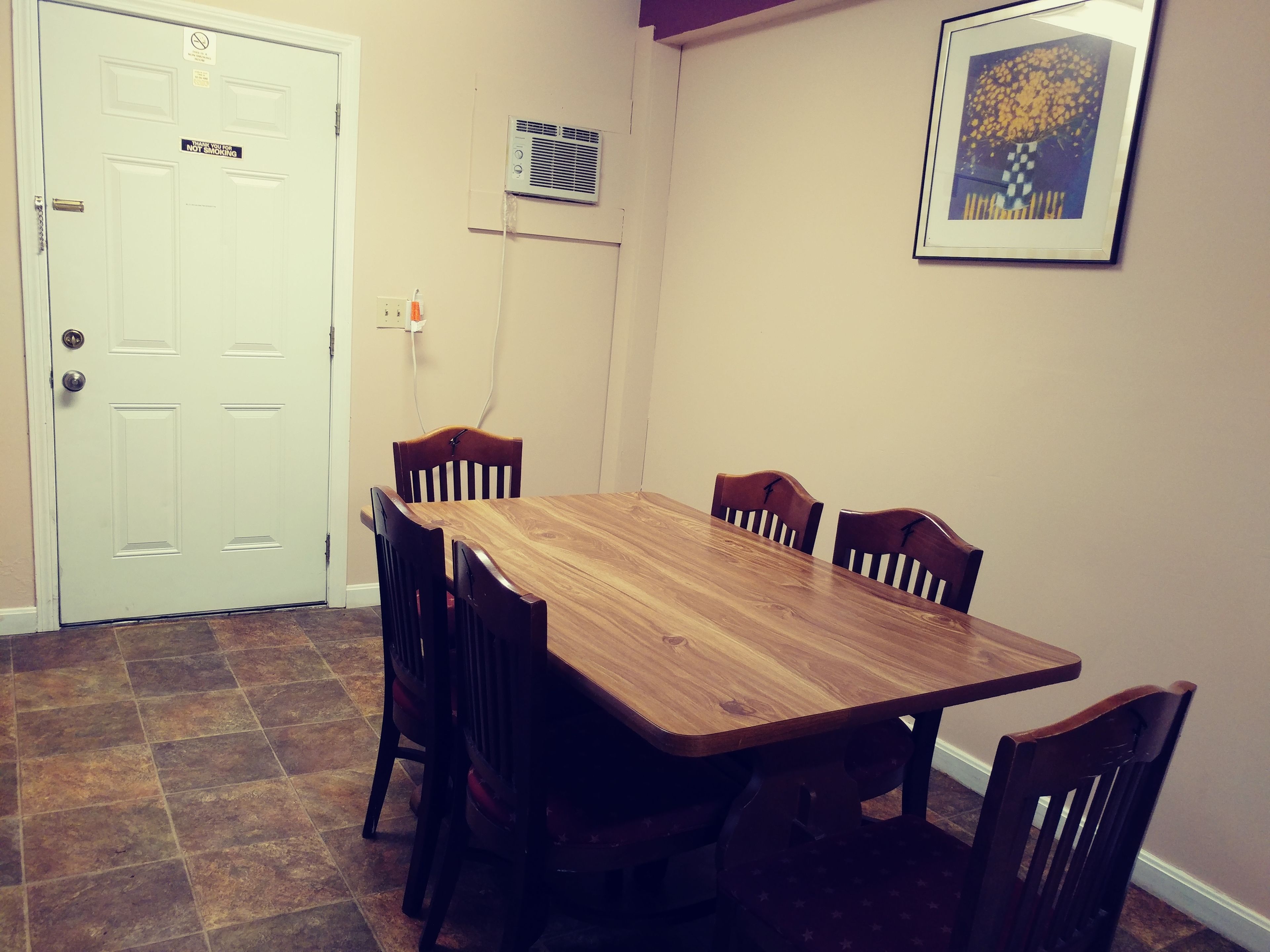 A dining area with a wooden table and chairs, a white door with a 'No Smoking' sign, a wall-mounted air conditioner, and a framed picture.