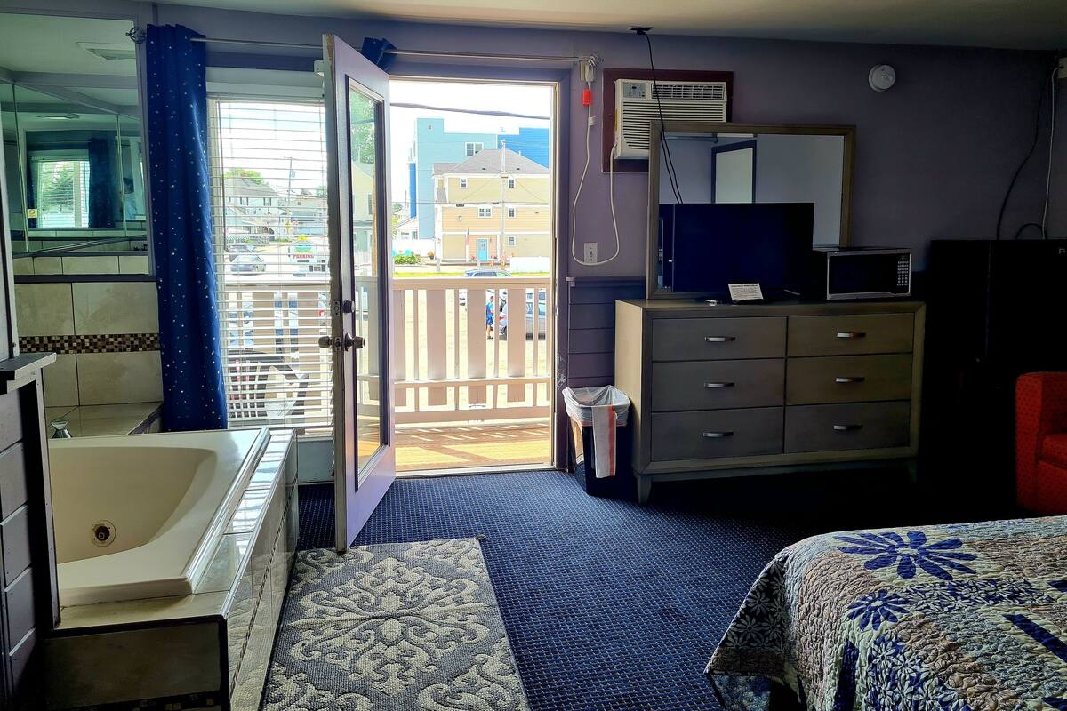 A hotel room with a bathtub, a wardrobe, a television, and a balcony with a view of the city.