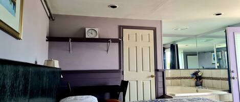 A bedroom with a bed, a nightstand, a clock, a door, and a bathroom visible through an open door.
