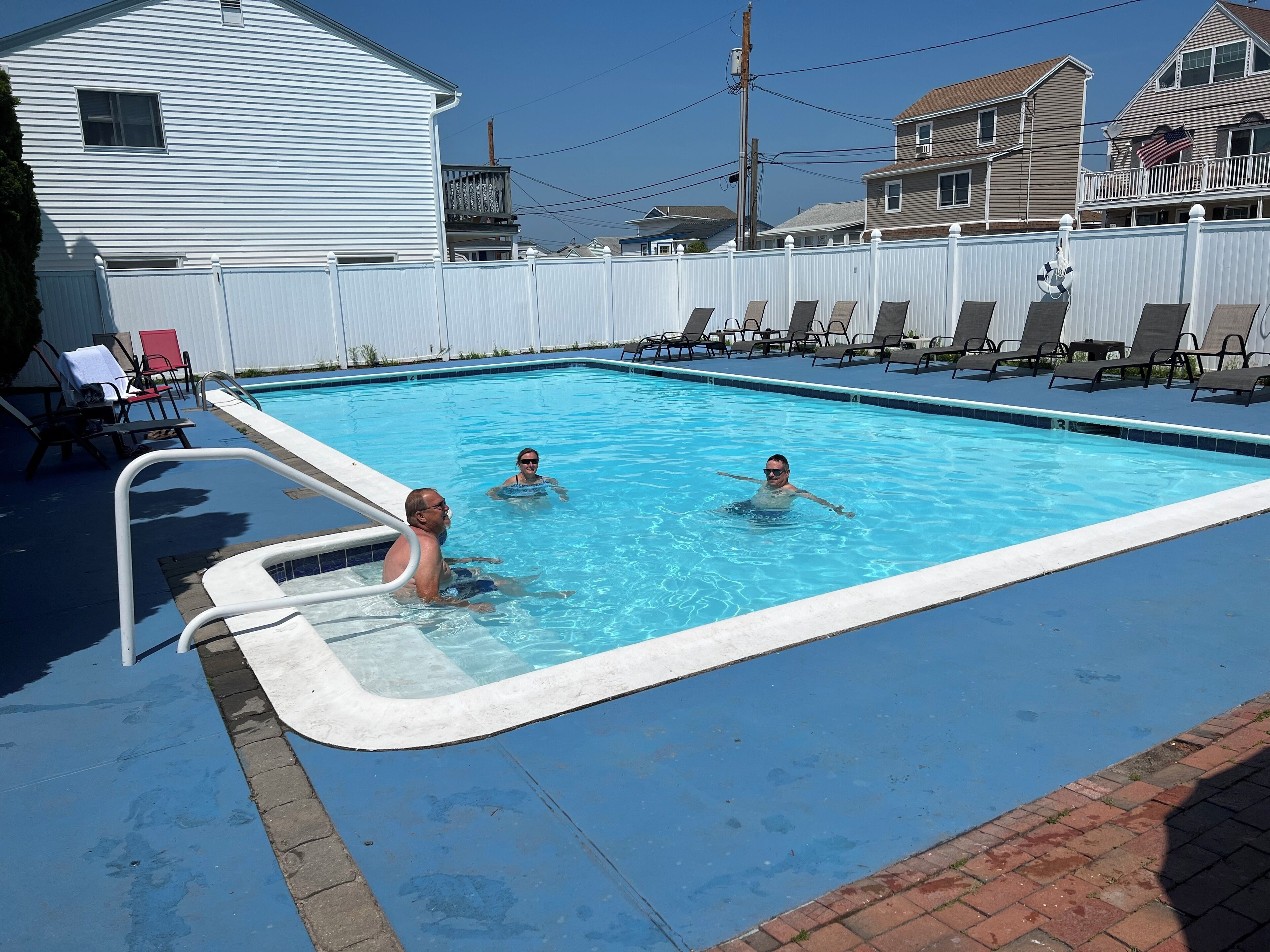 A swimming pool with three people in the water, surrounded by lounge chairs and a white fence.