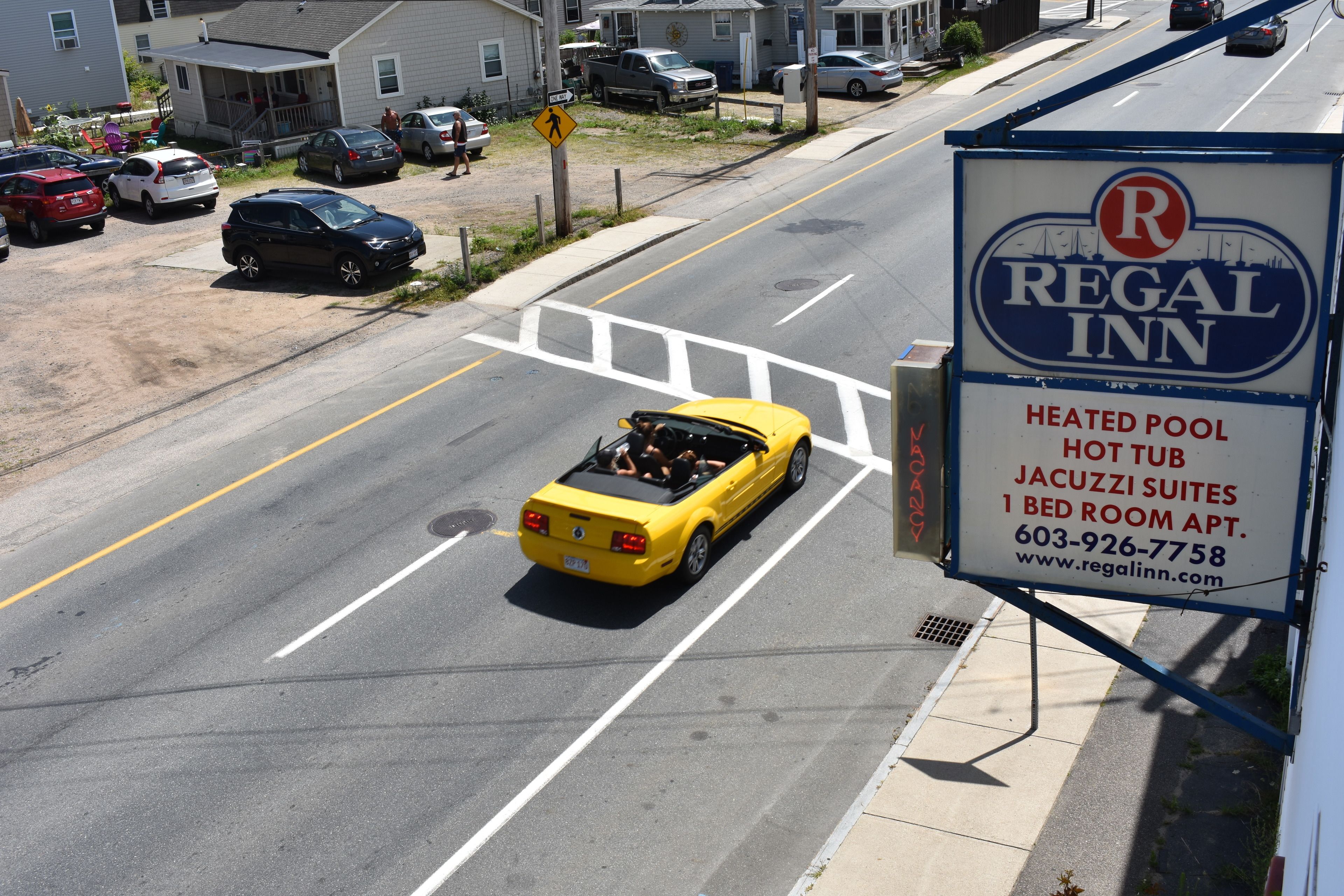 A yellow convertible driving on a road with a sign for Regal Inn advertising heated pool, hot tub, Jacuzzi suites, and 1 bed room apartment.