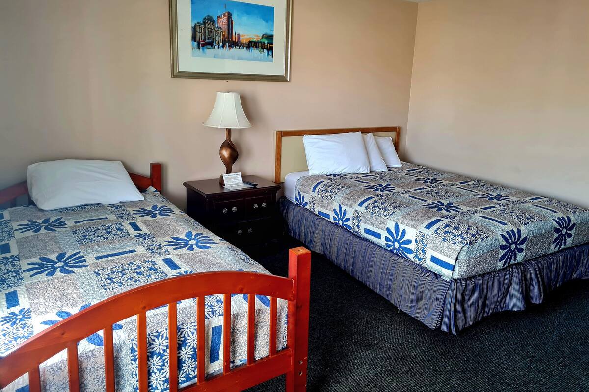 A hotel room with two beds, a nightstand with a lamp, and a framed cityscape painting on the wall.
