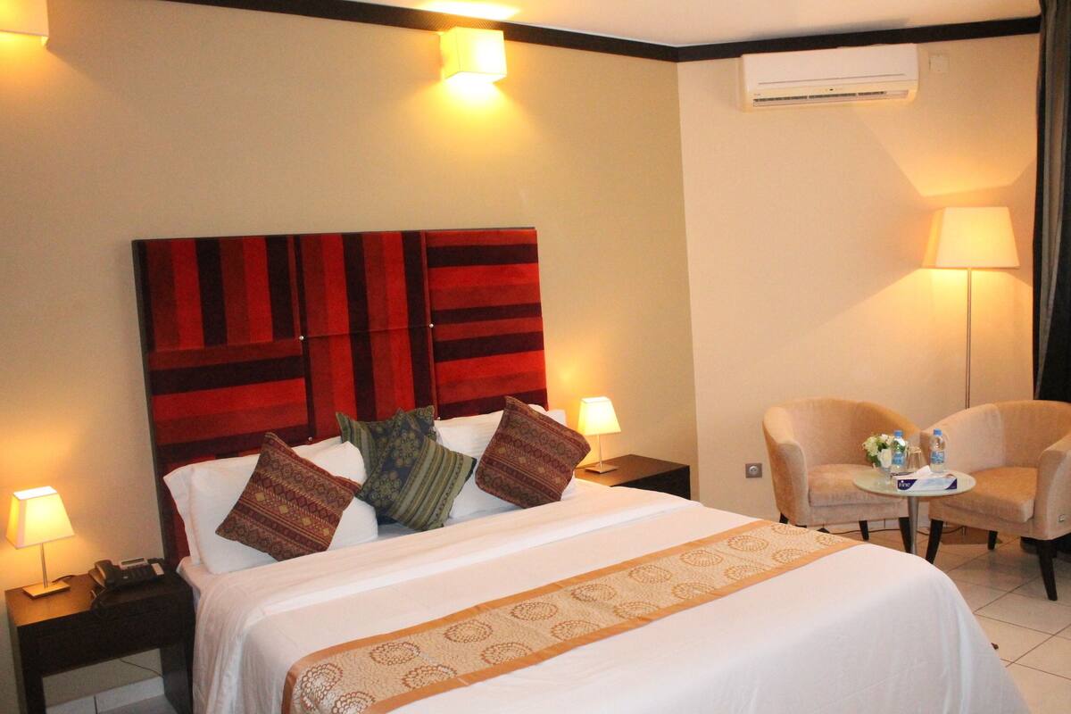Ein Hotelzimmer mit einem großen Bett, einem hölzernen Kopfteil, zwei Nachttischen mit Lampen, einem kleinen runden Tisch mit Stühlen und einer Wandklimaanlage.