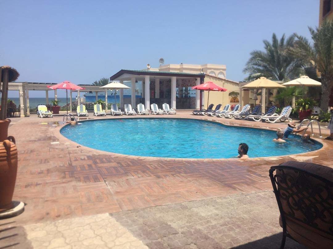 A swimming pool with lounge chairs and umbrellas, surrounded by palm trees and a building with columns.