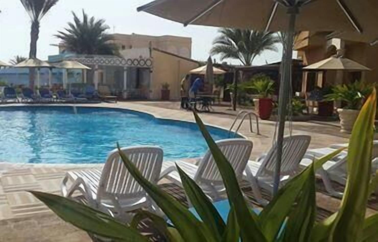 A swimming pool with lounge chairs and umbrellas, surrounded by palm trees and buildings.