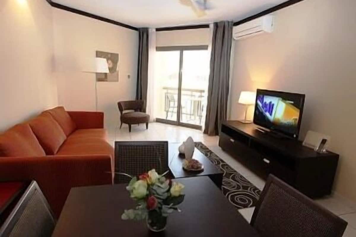 Ein Hotelzimmer mit einer Couch, einem Esstisch mit Blumenvase, einem Fernseher und einem Deckenventilator.