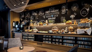 Een café met een prikbord waarop verschillende berichten en tekeningen staan, een koffieapparaat en diverse gebakjes en lekkernijen.