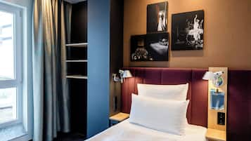 A hotel room with a bed, a nightstand, a lamp, a wall-mounted shelf, and a window with curtains.