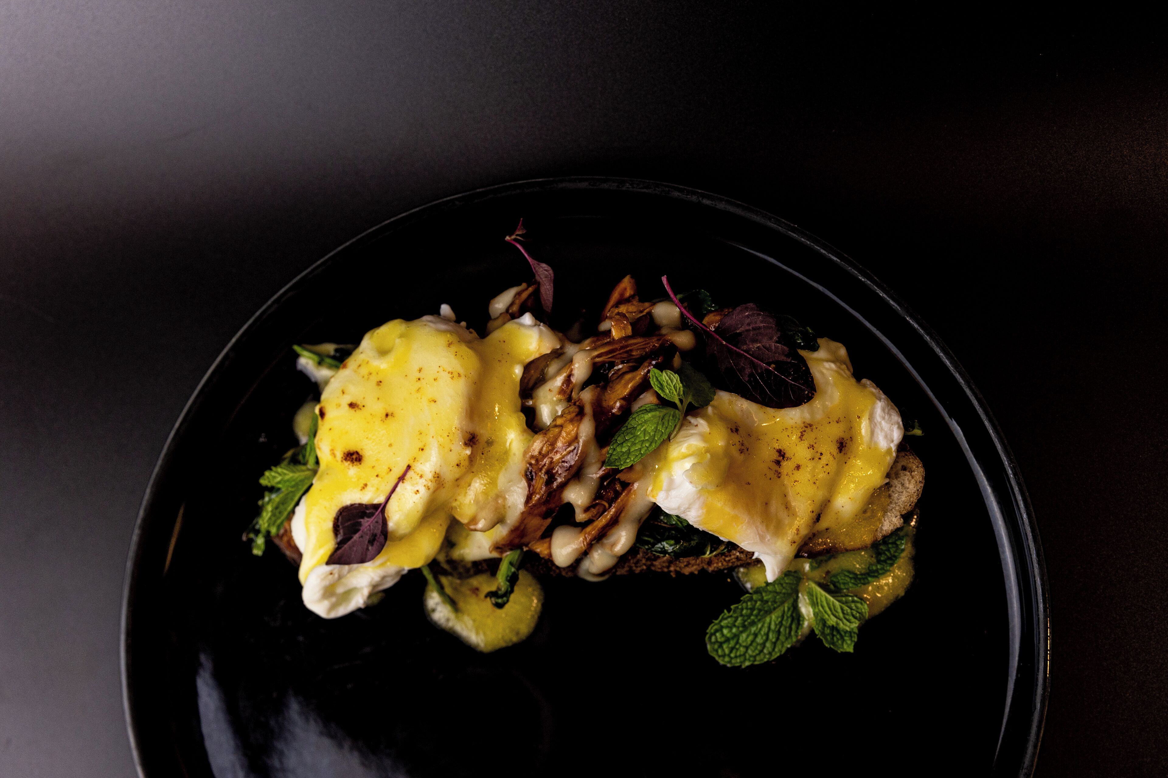 A black plate with a dish of eggs benedict, garnished with herbs and a slice of pineapple.