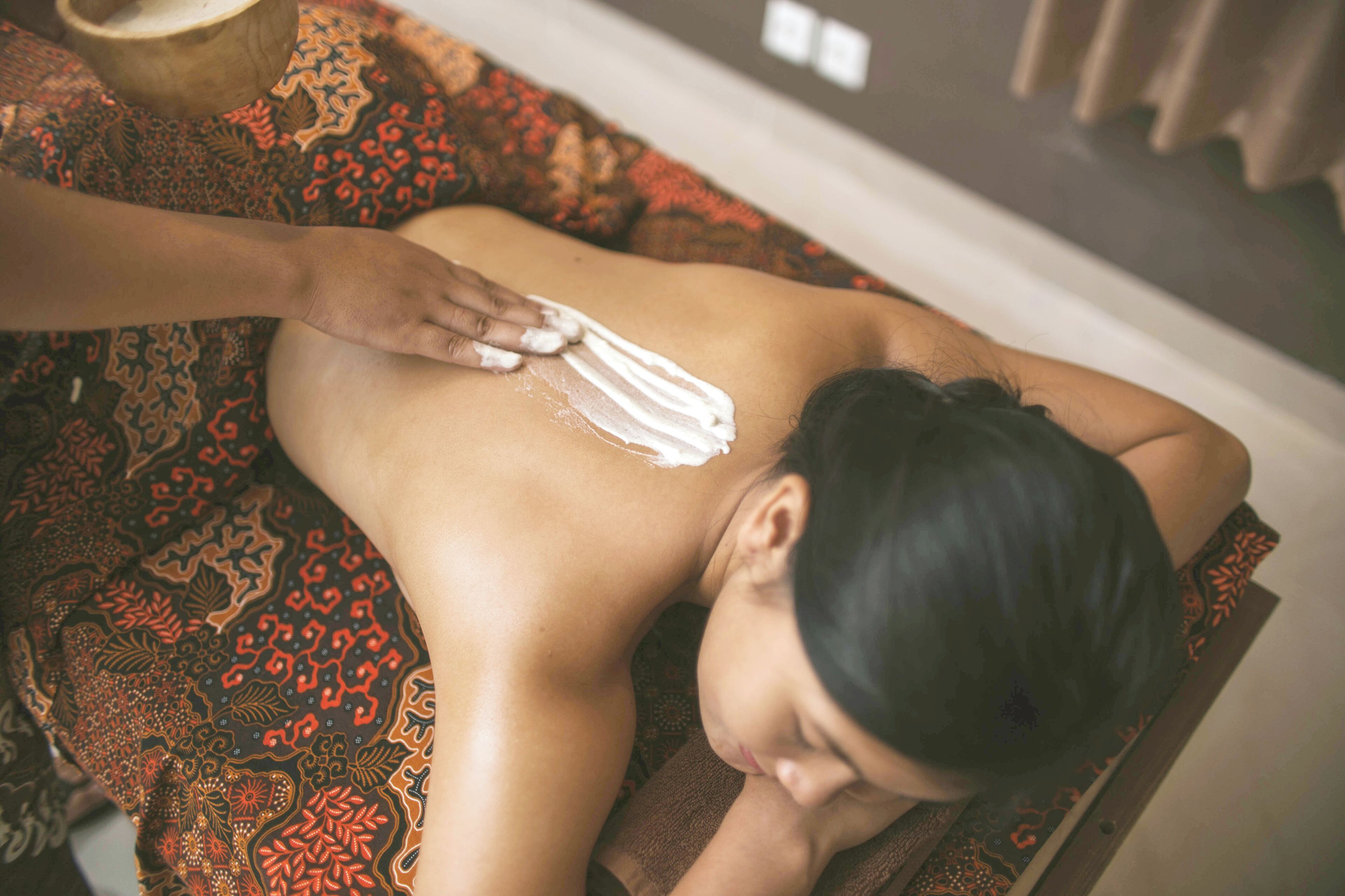 A person receiving a back massage in a room with patterned carpet and a wooden chair.