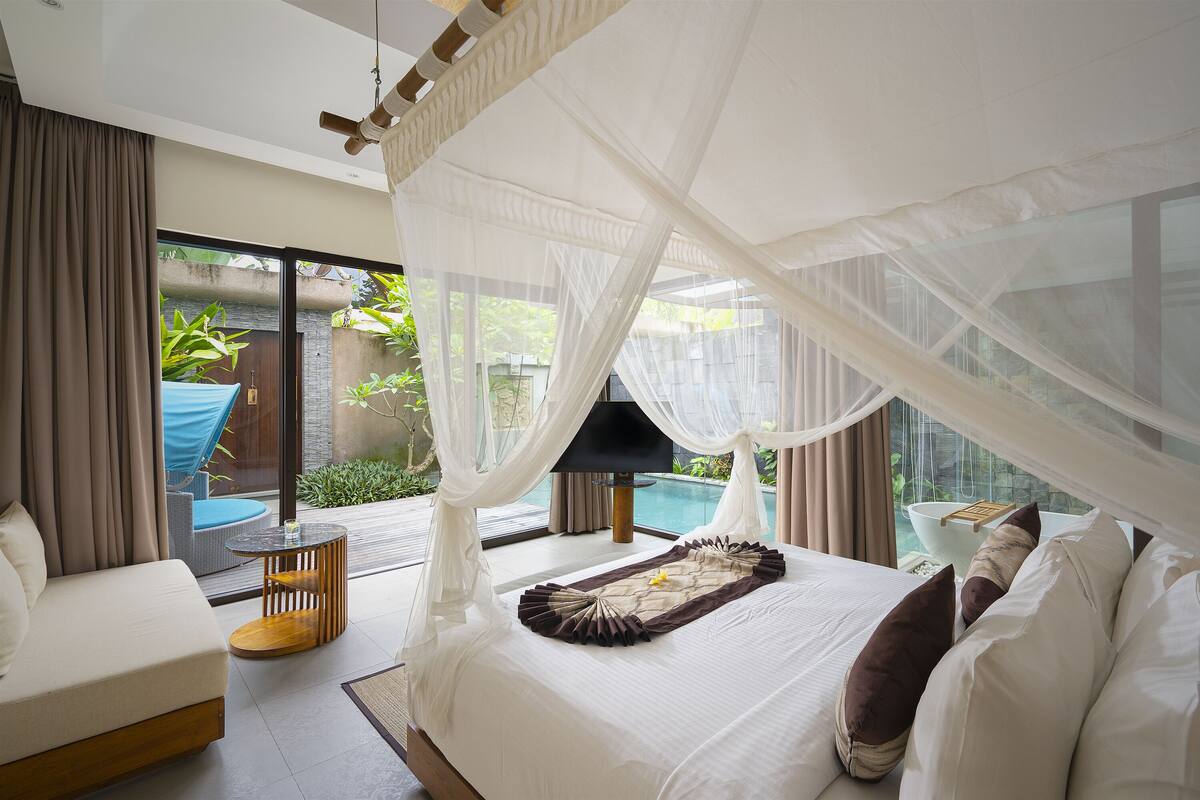 A bedroom with a bed, a sofa, a small table, and a view of a swimming pool and greenery.