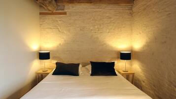 Un dormitorio con techo de madera, paredes de piedra, una cama con ropa blanca y dos lámparas de noche.