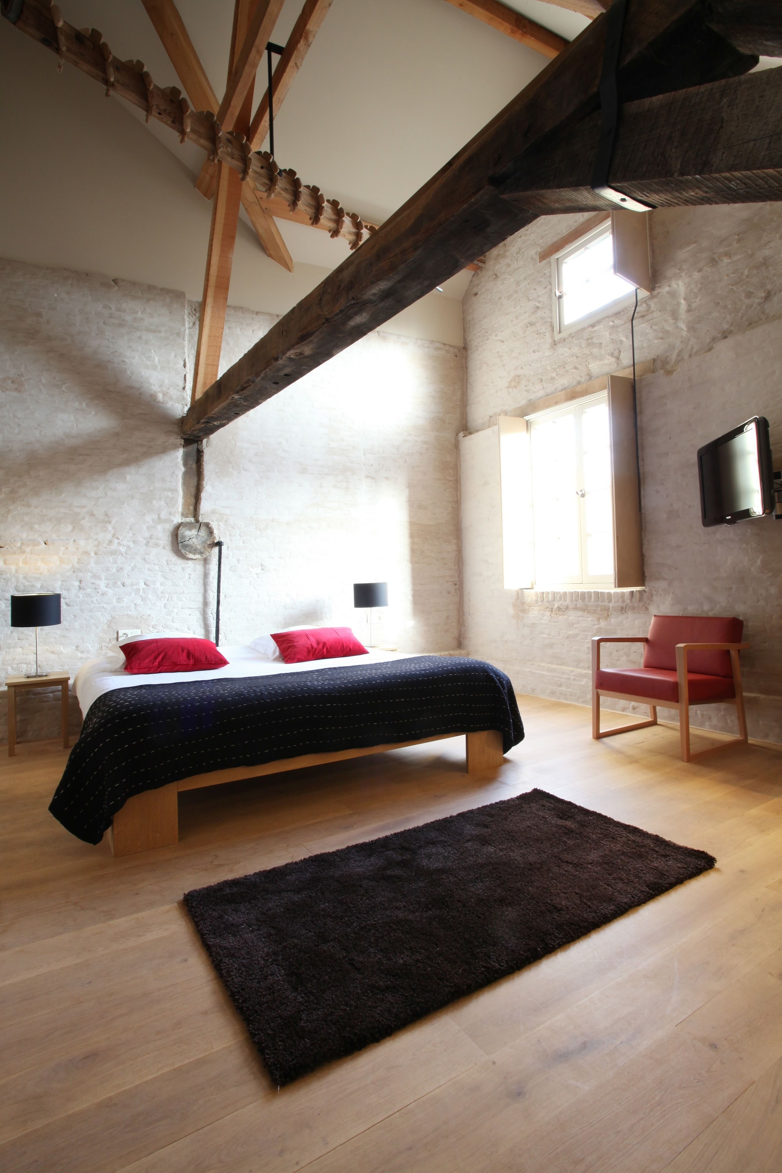 A bedroom with a wooden ceiling, a bed with red pillows, a dark rug, and a red chair.