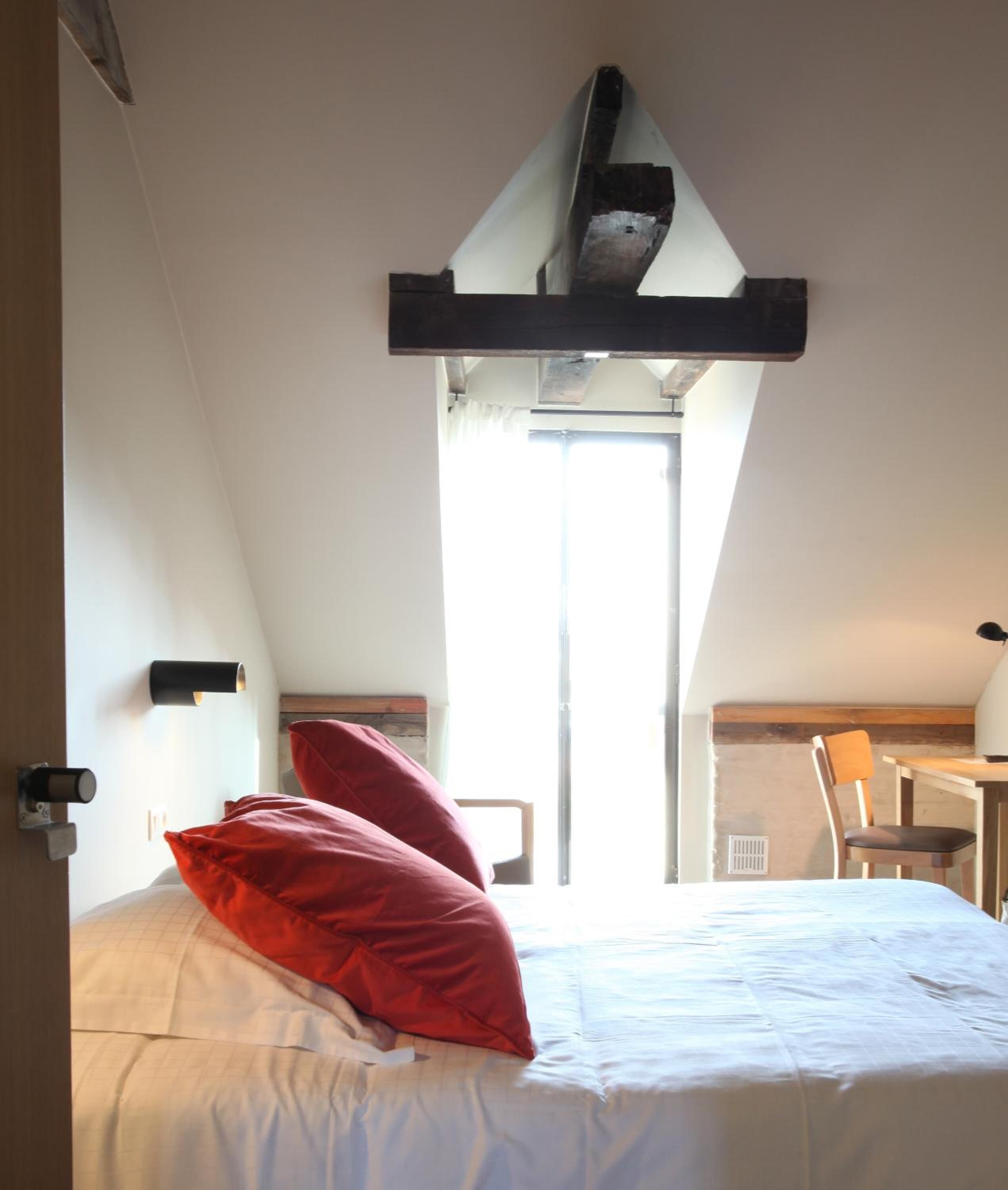 A bedroom with a bed, a red pillow, a wooden headboard, a desk, and a chair.