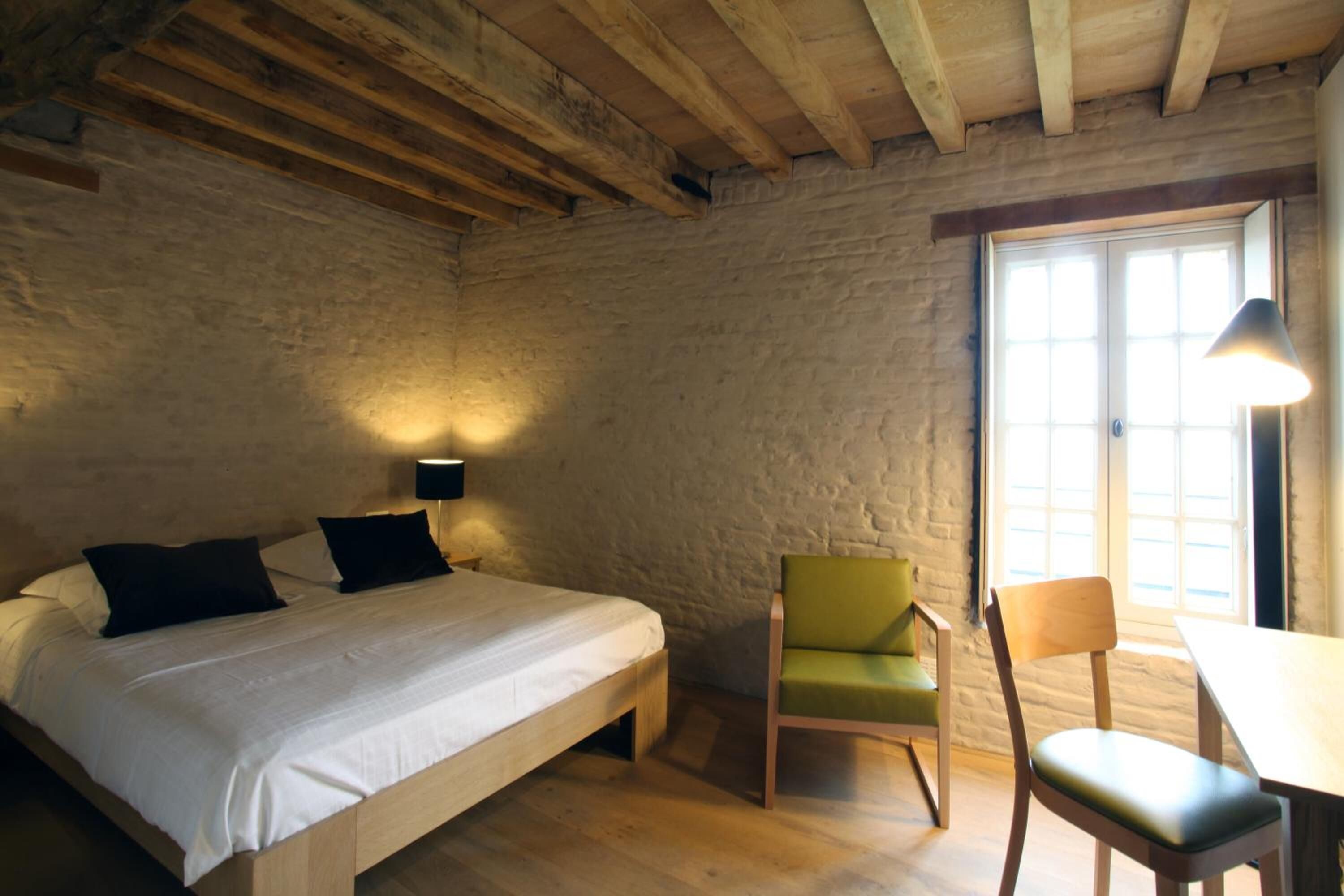 A bedroom with a wooden bed, a green chair, a wooden table, and a window with natural light.