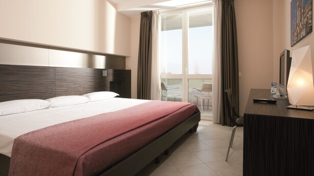 Een hotelkamer met een bed, een bureau met lamp, een stoel, en uitzicht op het landschap door een groot raam.
