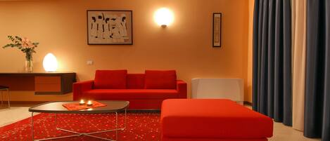 A modern living room with a red sofa, a small round table, and a red ottoman. There is a decorative vase with flowers on a side table, a wall-mounted light fixture, and a framed artwork above the sofa.