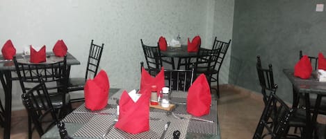 A dining area with tables set for four, each with a red napkin, black chairs, and a picture on the wall.