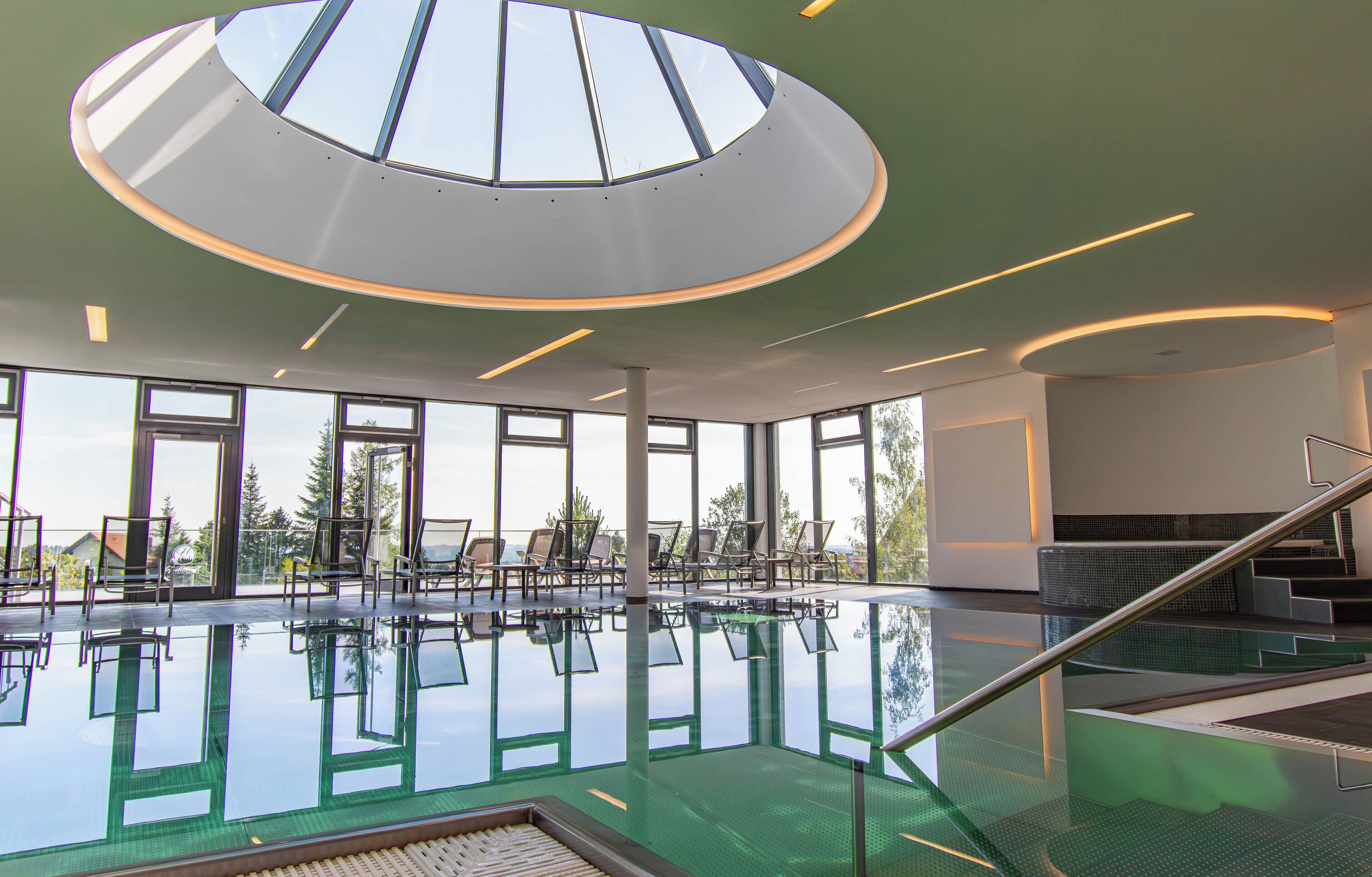 A modern indoor swimming pool with clear glass walls, a skylight, and a sunlit interior.