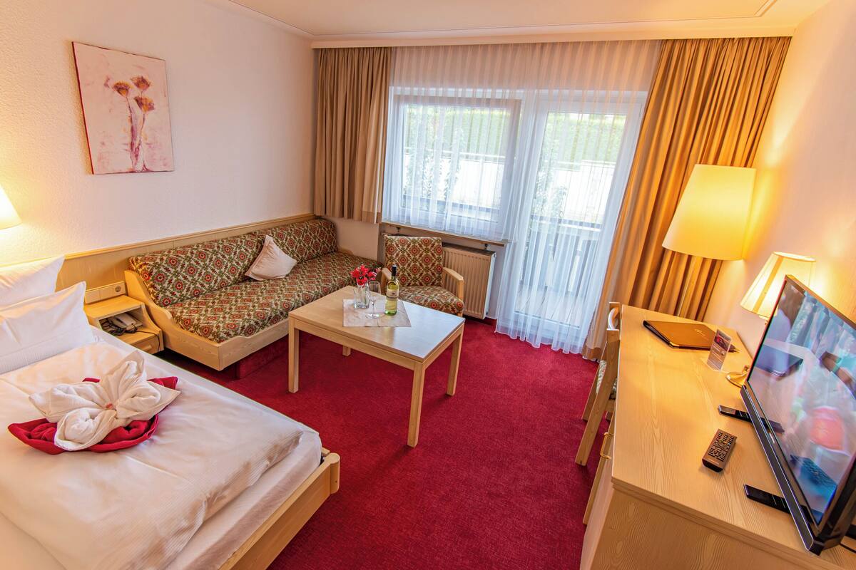 Quarto de hotel com cama, sofá, mesinha com garrafa e copos, televisão e janela com cortinas.