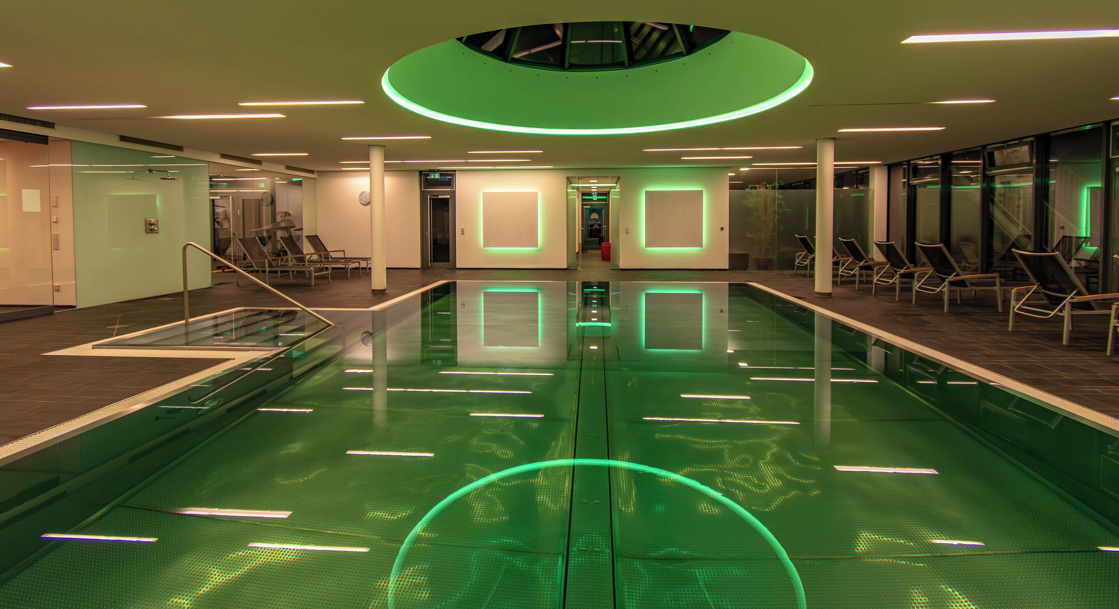 Piscina interior moderna com barreira de vidro, iluminação ambiente esverdeada e áreas de estar.